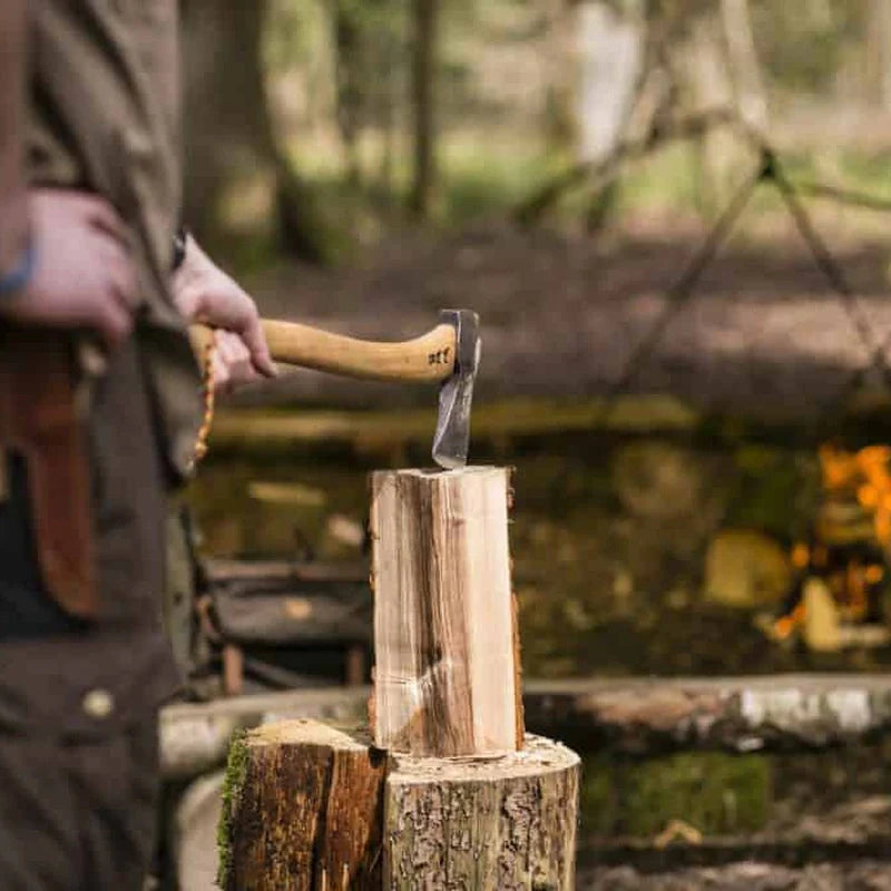 Bushcraft axe with wooden handle embedded in a split log beside a campfire in the woods